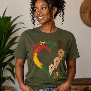May include: Olive green t-shirt featuring a heart-shaped design in the colors of the Pan-African flag, with the word "Juneteenth" and the year "1865" printed on it. The design also includes raised fists. The shirt is worn by a person.