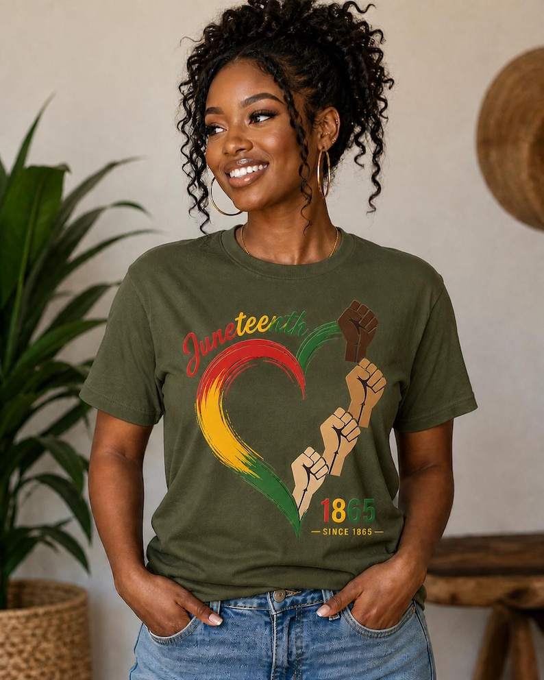 May include: Olive green t-shirt featuring a heart-shaped design in the colors of the Pan-African flag, with the word "Juneteenth" and the year "1865" printed on it. The design also includes raised fists. The shirt is worn by a person.