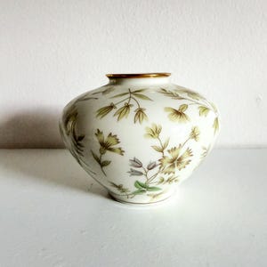 May include: A round, white ceramic vase featuring a delicate botanical pattern of light green and muted yellow flowers and leaves. The vase has a smooth, glossy finish and a thin, metallic gold rim around the circular opening at the top.