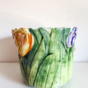 May include: A ceramic planter with a tulip design. The planter is painted with green leaves and features orange, yellow, and purple tulip blooms. The planter has a textured surface and a scalloped rim.