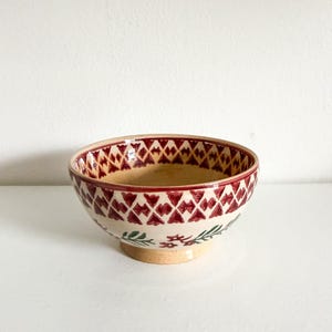 May include: A ceramic bowl with a cream-colored base and a red and cream diamond pattern around the rim. The bowl has green floral accents near the base. The interior of the bowl is red.