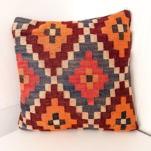 May include: A square throw pillow featuring a woven geometric pattern in shades of orange, deep red, slate blue, and cream. The design consists of repeating diamond and stepped shapes across the entire surface of the textile cover.