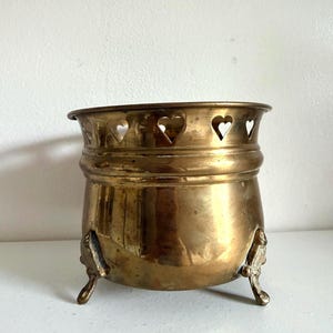 May include: A polished brass planter featuring a rounded body and three ornate feet. The upper rim is decorated with a series of cut-out heart shapes, providing a decorative border around the top edge of the metallic vessel.