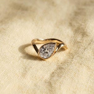 Könnte beinhalten: Ein Goldring mit einem einzelnen birnenförmigen Diamanten in einer Zargenfassung. Die Ringschiene hat ein glattes, wellenförmiges Design, das sich um den zentralen Stein schwingt und eine fließende Form aus poliertem Gelbgold bildet.