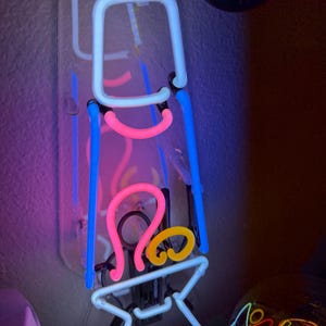 Puede incluir: Un letrero de neón con un respaldo transparente, con un cuadrado azul claro, líneas curvas rosas y un remolino amarillo. El letrero también tiene una base blanca y líneas verticales azules. El letrero está montado en una pared.