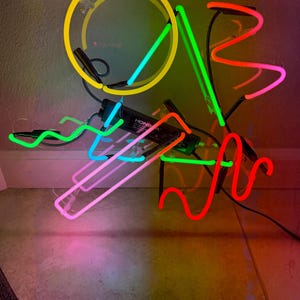 Könnte beinhalten: Eine farbenfrohe Neonlichtinstallation mit einem gelben Kreis und Röhren in Blau, Grün, Pink und Rot. Die Leuchte hat ein schwarzes Netzteil mit dem Text "HONGA". Die Neonlichter sind in abstrakten Formen angeordnet.
