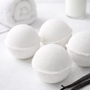 May include: Four white bath bombs with a textured surface are arranged on a white surface. A rolled white towel, a clear bottle, and two vanilla pods are also present. The image suggests a spa or self-care theme.