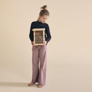 May include: A young person holds a wooden bird feeder filled with birdseed. The feeder is rectangular with a clear front panel. The person is wearing a navy blue long-sleeve shirt and mauve-colored pants.