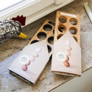 Puede incluir: Soportes de madera para huevos con seis recortes circulares, junto con embalajes con ilustraciones de huevos apilados y el texto "DAS EIERHUS". Los soportes son de color marrón claro, con el texto "Hanehus" visible. El embalaje dice "Design trifft Natur - handmade in Österreich."