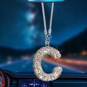May include: A sparkling, iridescent letter "C" car charm. The charm is encrusted with numerous, small, colorful crystals. It hangs from a silver chain with a lobster clasp. The background is a blurred image of a car interior and a night scene.