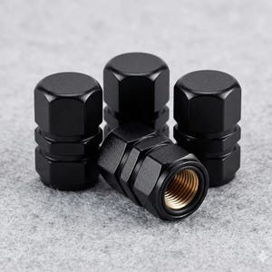 Puede incluir: Cuatro tapones negros para válvulas de neumáticos con diseño hexagonal. Los tapones tienen un acabado mate y un interior dorado. Los tapones están dispuestos sobre una superficie gris.