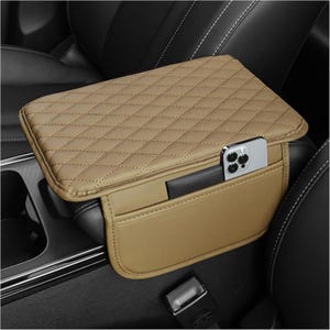 Puede incluir: Funda de reposabrazos para coche de color beige con diseño acolchado. La funda tiene un bolsillo con un smartphone y una pequeña cartera negra. Diseñada para adaptarse al reposabrazos central de un coche.