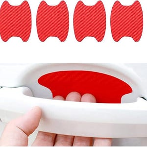 May include: Four red carbon fiber textured car door handle protectors. The protectors are shaped to fit the door handle recess and are shown with a hand using the door handle.