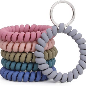 Könnte beinhalten: Ein Stapel bunter Spiral-Armband-Schlüsselanhänger in Rosa, Pfirsich, Olivgrün, Türkis und Blau. Ein grauer Schlüsselanhänger ist oben befestigt. Die Schlüsselanhänger bestehen aus einem weichen, flexiblen Material.