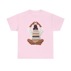 May include: Pale pink t-shirt with an illustration of a person sitting cross-legged, holding a stack of books. The person's arms are raised, forming a heart shape above their head. The person is wearing a white shirt.