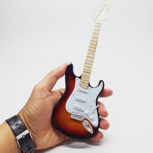 May include: Miniature electric guitar with a sunburst finish, white pickguard, and chrome hardware. The guitar has a light-colored neck and headstock. A small-scale replica of a classic instrument.