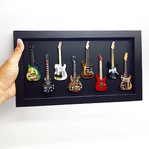 Może przedstawiać: Czarna oprawiona ekspozycja z ośmioma miniaturowymi gitarami elektrycznymi. Gitary są w różnych kolorach, w tym czerwonym, czarnym i brązowym, ze szczegółowymi wzorami. Ramka jest trzymana przez dłoń na białym tle.
