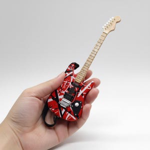 May include: Miniature red and white electric guitar with black accents. The guitar has a wooden neck and headstock, and is held in a hand. The guitar is a replica of a famous model.