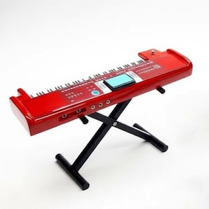 May include: A bright red electronic keyboard with a black stand. The keyboard has a small blue screen and multiple buttons and keys. The stand is a black, X-shaped design, providing support. The keyboard is set against a white background.