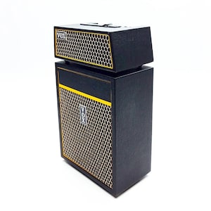 Könnte beinhalten: Schwarzer und goldfarbener Miniatur-Gitarrenverstärker mit dem sichtbaren Markennamen "VOX". Der Verstärker hat eine rechteckige Form mit einem Lautsprechergitter und gelben Zierleisten. Der obere Teil ist eine separate Einheit.