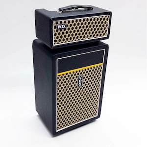 Könnte beinhalten: Ein schwarzes und goldenes VOX-Verstärker-Set. Das Set enthält einen kleinen Verstärkerkopf mit Griff und eine größere Lautsprecherbox. Beide Teile haben ein goldfarbenes Gitter und das VOX-Logo.