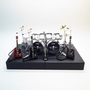 Op de afbeelding: Miniatuur rockband set op een zwart podium. De set bevat een rode gitaar, twee zwarte gitaren, een drumstel met schedelafbeeldingen, bekkens en een microfoon. De bassdrum van het drumstel heeft een schedelafbeelding.