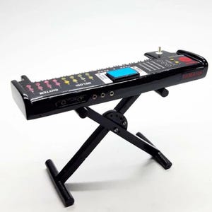 Puede incluir: Un teclado Yamaha negro en miniatura con una pantalla azul y un soporte. El teclado tiene las palabras "RHYTHM" y "MELODI" impresas. El soporte es negro y tiene un diseño en forma de X.