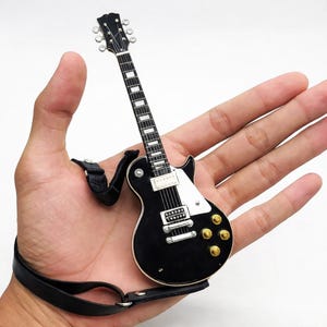 May include: Miniature black electric guitar with a white pickguard and gold knobs. The guitar has a black strap and silver tuning pegs. The fretboard is white with black frets. A detailed replica of a classic instrument.