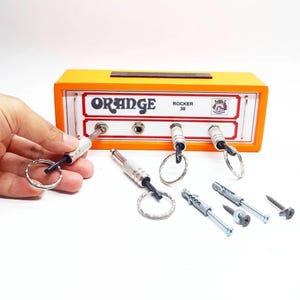 May include: Orange Rocker 30 key holder with three keychains, screws, and wall anchors. The key holder is orange with the text "ORANGE ROCKER 30". The keychains are silver and black.
