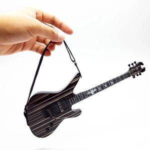 Könnte beinhalten: Miniatur-E-Gitarre mit einem schwarz-weißen Streifendesign. Die Gitarre hat einen schwarzen Gurt und wird vor einem weißen Hintergrund gehalten. Das Griffbrett der Gitarre hat dekorative Elemente.
