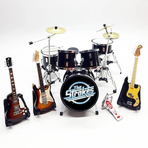 Op de afbeelding: Miniatuur zwart drumstel met bekkens en verschillende gitaren. De grote trommel heeft de tekst "The Strokes" in een wit en blauw ontwerp. De gitaren zijn bruin, zwart en geel.