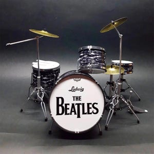 Op de afbeelding: Miniatuur drumstel met een witte basdrum met het "The Beatles" logo. De set bevat zwart-wit patronen drums, goudkleurige bekkens en zilveren standaards. Een voetpedaal is ook zichtbaar.