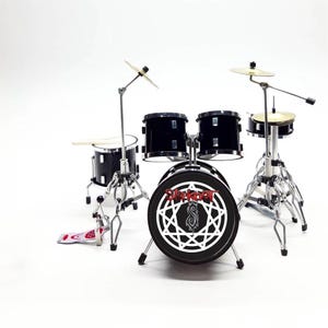 Puede incluir: Una batería en miniatura de color negro que presenta un bombo con el logotipo de Slipknot y un símbolo de nonagrama en blanco en el parche frontal. El set incluye dos toms aéreos, un tom base, platillos metálicos y un pedal blanco y rojo.