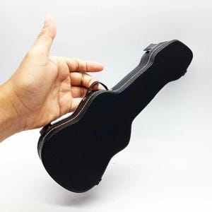 Może przedstawiać: Czarny futerał na gitarę z białymi przeszyciami i uchwytem. Futerał ma kształt małej gitary i wydaje się być wykonany ze skóry lub podobnego materiału. Futerał jest trzymany na białym tle.