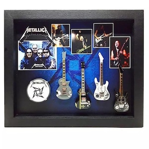 Może przedstawiać: Oprawiona ekspozycja z zespołem Metallica. Ekspozycja zawiera zdjęcia zespołu, logo perkusji i miniaturowe repliki gitar. Tło jest niebieskie ze stylizowanym wzorem. Rama jest czarna.
