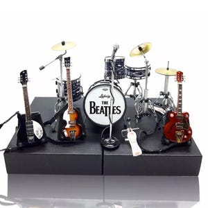 Può includere: Set in miniatura della band dei Beatles con batteria, chitarre e un'asta per microfono. Il nome della band è stampato sul tamburo. Le chitarre sono nere, marroni e rosse. Il set è esposto su una piattaforma nera.