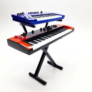 May include: A miniature, two-tiered keyboard setup. The top keyboard is blue, and the bottom one is red. Both have white keys and black accents. The keyboards are mounted on a black stand, creating a layered effect. The setup is designed to resemble a professional musical instrument.