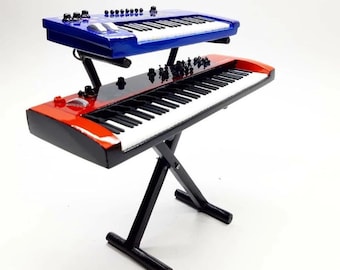 May include: A miniature, two-tiered keyboard setup. The top keyboard is blue, and the bottom one is red. Both have white keys and black accents. The keyboards are mounted on a black stand, creating a layered effect. The setup is designed to resemble a professional musical instrument.