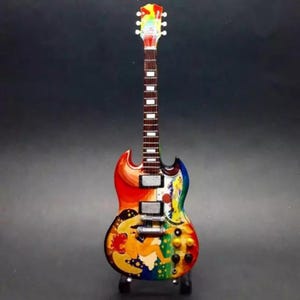 Op de afbeelding: Een kleurrijke miniatuur elektrische gitaar met een regenboogafwerking. De body heeft een levendig ontwerp met een vrouw, maan en sterren. De gitaar heeft een witte hals met frets en een regenboog kop.