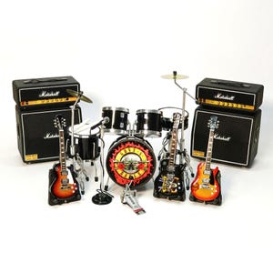 Peut inclure: Ensemble miniature du groupe Guns N' Roses comprenant une batterie, deux amplificateurs Marshall et trois guitares. La grosse caisse de la batterie porte un logo. Les guitares sont orange, noires et marron. Les amplificateurs sont noirs avec du texte blanc.