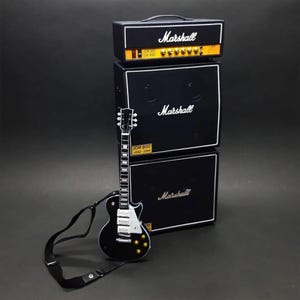 Könnte beinhalten: Eine Miniatur-E-Gitarre in Schwarz mit einem schwarzen Gurt liegt vor einem Stapel schwarzer Marshall-Verstärker. Die Verstärker haben das Marshall-Logo in Weiß und gelben Akzenten. Die Gitarre hat silberne Hardware.