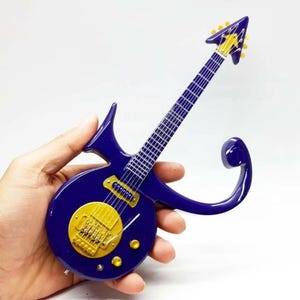 May include: A miniature electric guitar in a vibrant purple and gold color scheme. The guitar features a unique, stylized design with a round body and a distinctive headstock. The gold-colored details include the pickups and tuning pegs.