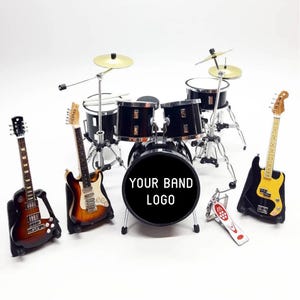 Può includere: Set di strumenti musicali in miniatura con una batteria con piatti e tre chitarre. La grancassa ha il testo "YOUR BAND LOGO". Gli strumenti sono neri, marroni e gialli.