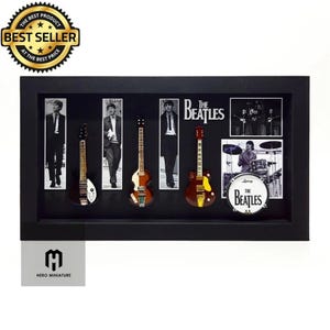 May include: A framed display featuring miniature guitars and images of The Beatles. The black frame holds five guitars, band photos, and a drum set. The words "THE BEATLES" and "BEST SELLER" are visible.