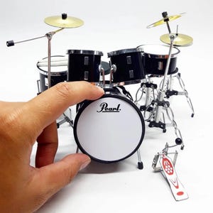 May include: Miniature black and silver drum set with a white bass drum head featuring the "Pearl" logo. The set includes cymbals, drum pads, and a bass drum pedal. The drum set is a small scale model.