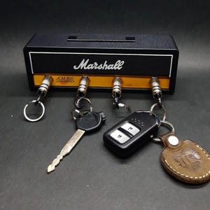 May include: Black key holder shaped like a Marshall amplifier with the brand name in white. The key holder has four key hooks and is holding keys and key fobs. The key holder has a yellow stripe and the text "JCM 800 LEAD SERIES".