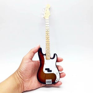 Puede incluir: Miniatura de bajo eléctrico con cuerpo marrón, blanco y negro. El mástil es de madera clara con trastes y clavijas. El instrumento se sostiene en una mano sobre un fondo blanco. El bajo es una pequeña réplica.