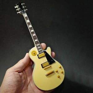 Puede incluir: Una guitarra eléctrica en miniatura de color crema con herrajes dorados y un golpeador negro. La guitarra tiene un diapasón blanco con incrustaciones rectangulares y un clavijero negro. El instrumento se sostiene en una mano sobre un fondo oscuro.
