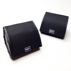 May include: Two black, rectangular audio speakers with a textured surface. Each speaker has a small, rectangular label that reads "TACT". The speakers are angled and appear to be made of a durable material.