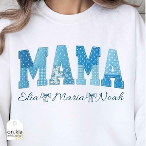 Könnte beinhalten: Weißes Sweatshirt mit dem Wort "MAMA" in blau geflickten Buchstaben. Darunter stehen die Namen "Elia Maria Noah" in Schreibschrift. Das Sweatshirt hat ein Etikett mit der Aufschrift "on.kia art&design".